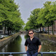 Auf dem Bild sieht man einen jungen Mann mit blauer Hose und schwarzem Hemd, der auf einer Brücke vor einem kleinen Fluss steht.