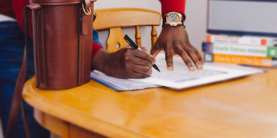 Man signs a document that lies on a table. Next to the document is a brown leather bag. 
