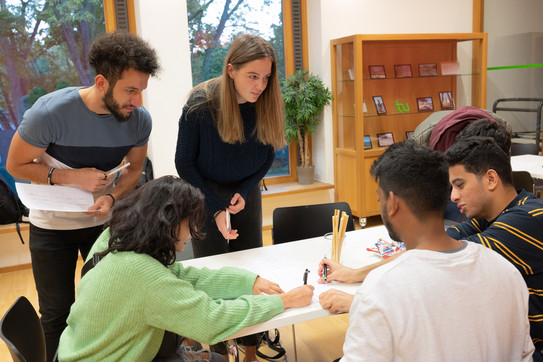 Sprachcafé_10 Einige internationale Studierende sitzen und stehen um einen Tisch. Sie sprechen miteinander.