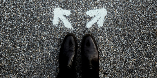 Praktikumsprogramme Füße in eleganten Schuhen stehen vor zwei Pfeilen auf dem Asphalt, die in unterschiedliche Richtungen zeiten.