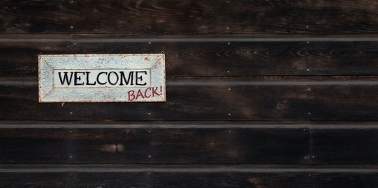 Welcome Back weißer "Welcome" Schild hängt am dunklen Holz. "Back" wurde handschriftlich hinzugefügt