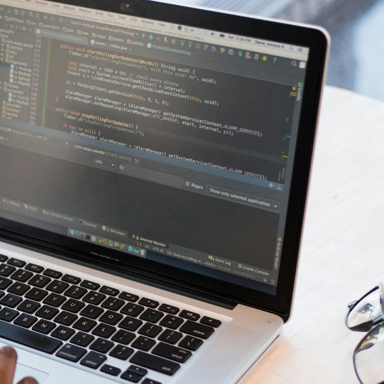 A woman is sitting at a round table in a café. On the table are a pair of glasses, a laptop, and a coffee mug. The woman is using her cell phone with her left hand and her laptop with her right hand.