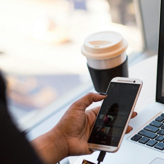 Eine Frau sitzt an einem runden Tisch eines Cafés. Auf dem Tisch liegen eine Brille, ein Laptop und ein Kaffeebecher. Mit der linken Hand bedient die Frau ihr Handy, mit der rechten Hand ihren Laptop.
