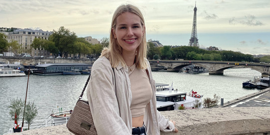 Profilbild Hier sieht man die Studentin, die an einem Vorsprung lehnt und in die Kamera lächelt. Im Hintergrund sieht man die Seine und den Eifelturm.