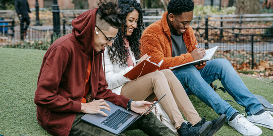 Campusleben_Vor der Bewerbung.jpg Drei junge Menschen sitzen auf dem Boden einer Parkwiese, die an einem sonnigen Tag grün ist. Eine Person verwendet einen Laptop, während die anderen in ihren Notizbüchern schreiben. Alle lachen und wirken in einem entspannten, gemeinsamen Lernmoment.