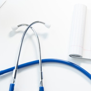 Auf einem weißen Tisch liegt ein blaues Stethoskop und ein aufgeschlagener Block. Darüber ist ein Teil eines Arms mit Armbanduhr zu sehen von einer Person, die einen blauen Kittel trägt.