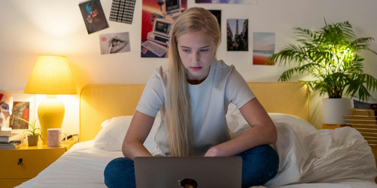 Eine Studierende sitzt auf einem Bett und blickt konzentriert auf den Laptop vor ihr