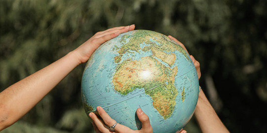 Two people reach for a globe from the left and right; only forearms are visible, background blurred green.