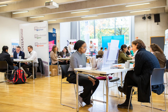 IKM_2023 In einem großen Raum stehen viele Tische, an denen Menschen Gespräche führen