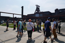 Menschen stehen zuhörend auf dem Gelände der Zeche Zollverein mit dem Förderturm im Hintergrund.