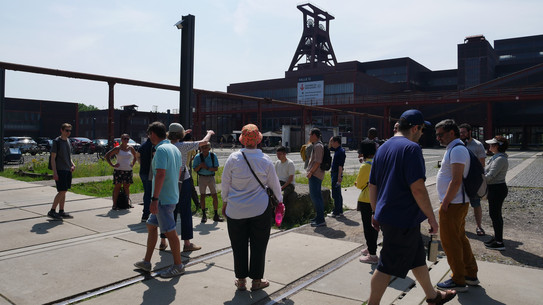 People are standing on the premises of Zeche Zollverein and listening, with the shaft tower in the background.