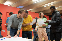 Sprachcafé_6 Some international students are standing next to a table and talking. They are showing each other on pieces of paper.