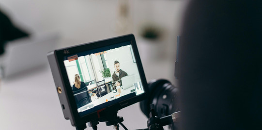 Der Kameramonitor zeigt eine Aufnahme von zwei Personen im Büro, eine sitzt mit dem Rücken zur Kamera, während die andere auf einem Laptop arbeitet.