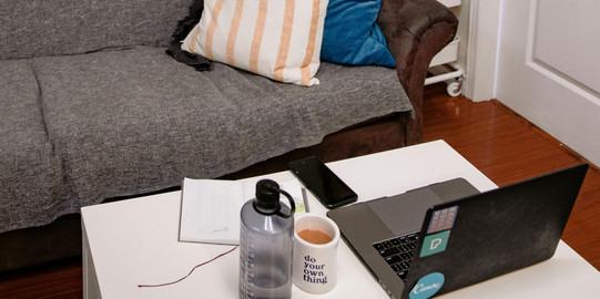 Ein gemütliches Wohnzimmer mit einem grauen Sofa, bunten Kissen und einem weißen Tisch. Auf dem Tisch stehen eine Wasserflasche, eine Tasse Kaffee und ein Laptop. An der Wand hängen Fotos und Bücherregale.