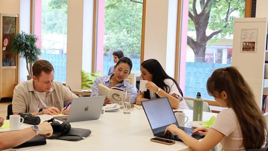 Four people at a table, two looking at a book, two at the computer