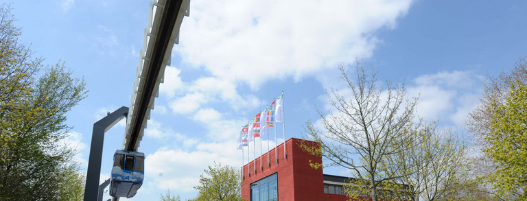 Internationales Begegnungszentrum mit H-Bahn Rotes Gebäude umgeben von Bäumen mit blauem Himmel und wenigen Wolken. Links im Bild die H-Bahn.